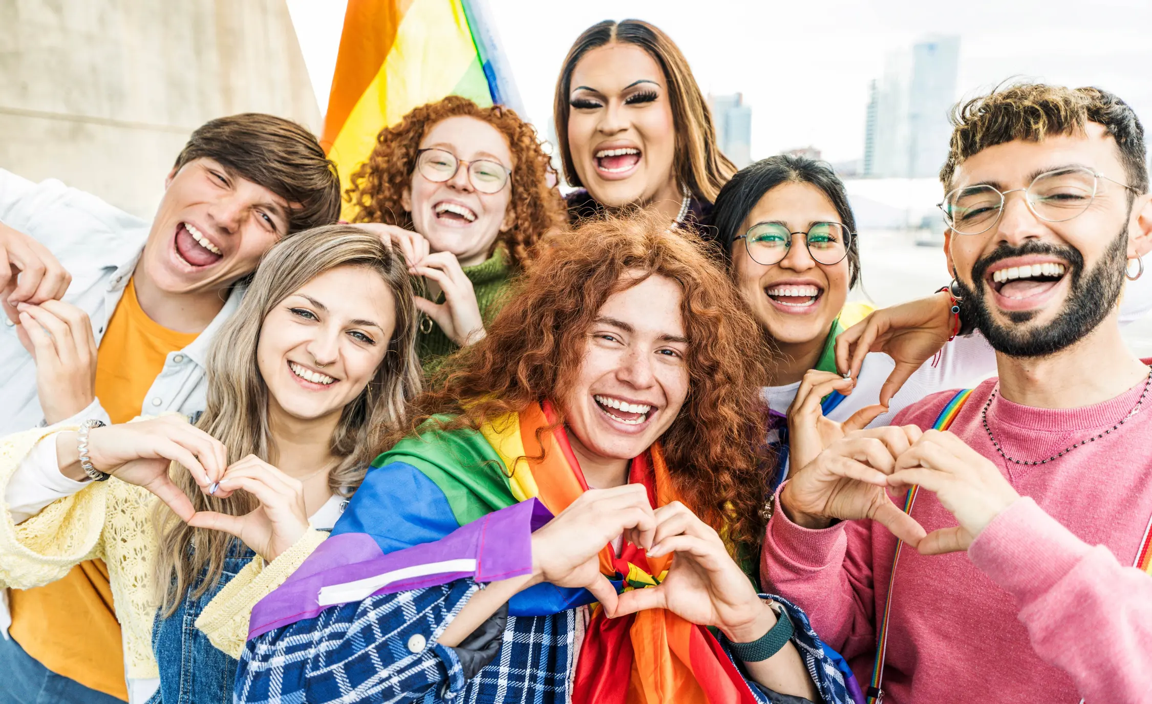 A diverse group of seven young people smiling and making heart shapes with their hands, several wearing or holding rainbow Pride flags